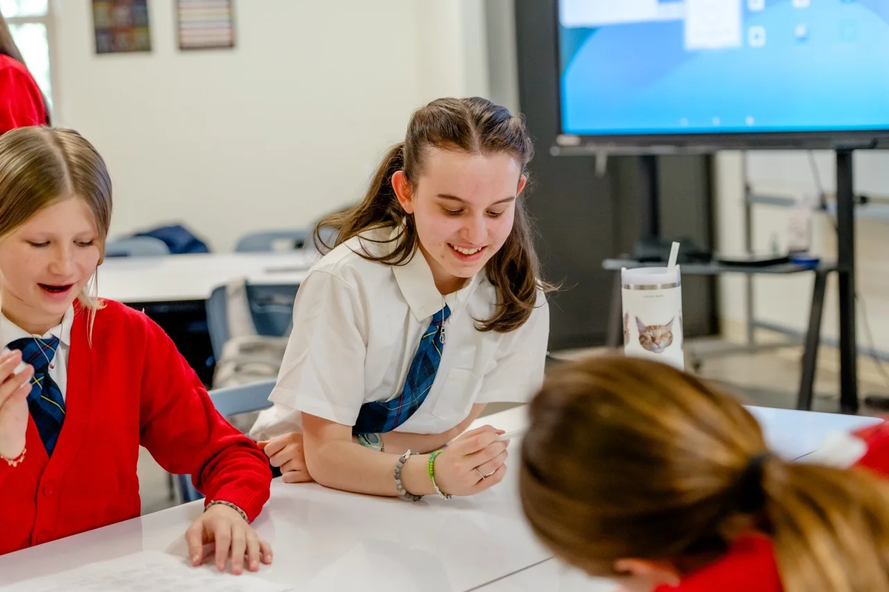Middle school students in white blouses and plaid ties smile and collaborate at a table during a lesson at The Texas Girls School, with a digital smartboard in the background.
