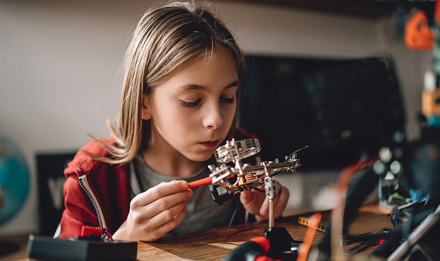 column-header-4 girl sitting at a desk or table using magnifying glass and tools to create something with gears