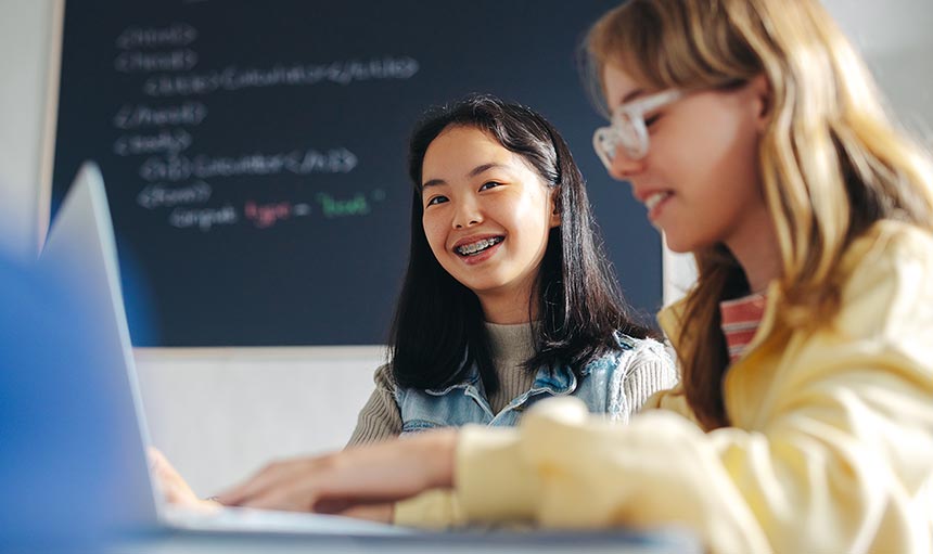 column-header two girls in class smiling. one girl has braces and brown hair and the other has blonde hair and glasses and is typing on a laptop