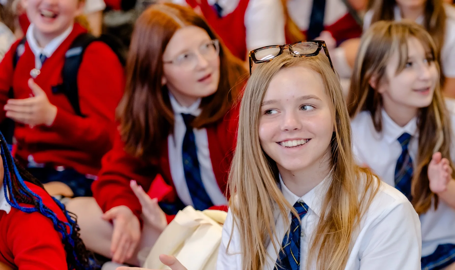 A group of Texas Girls School students in red cardigans and ties sit together, laughing during a school assembly.