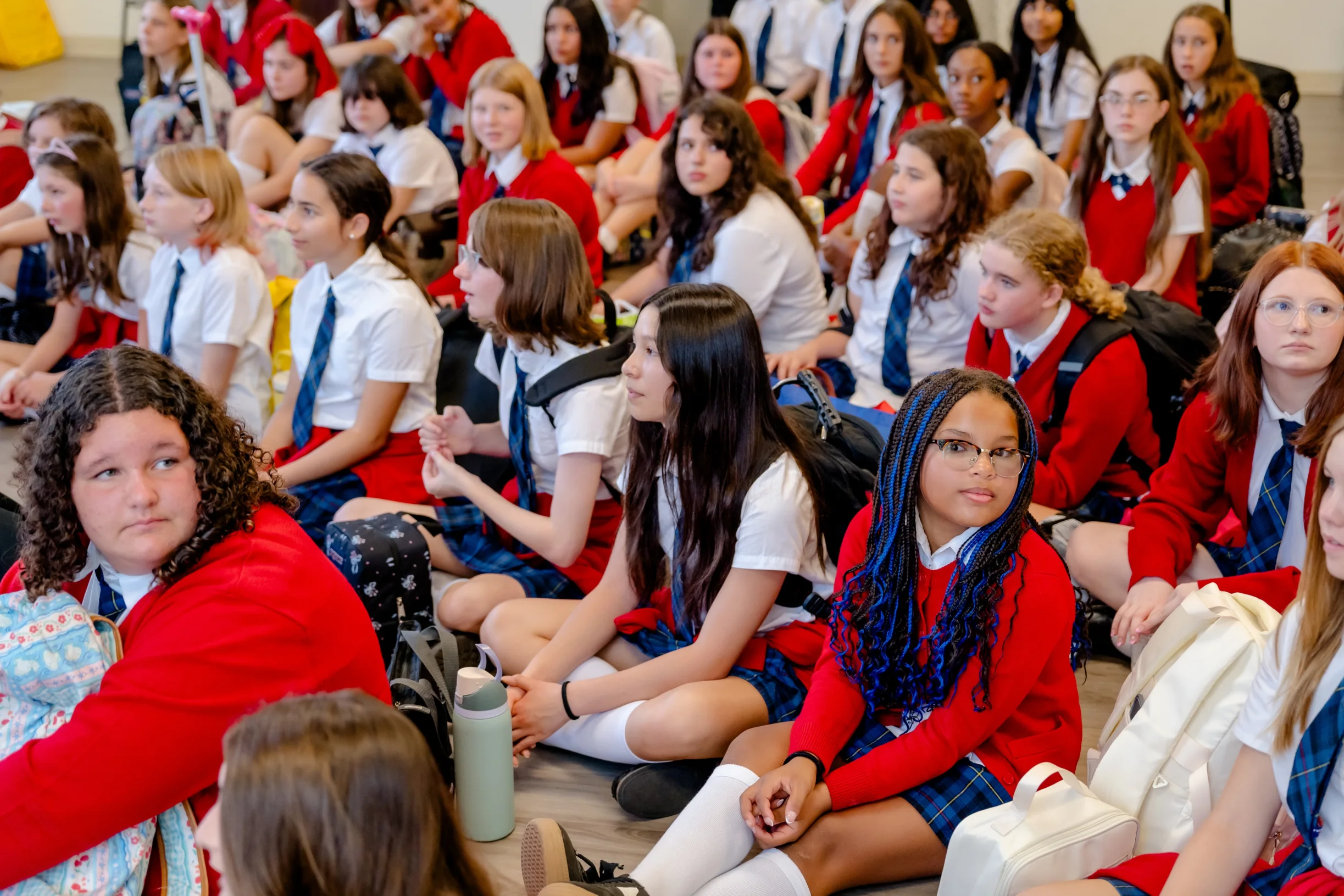 A large, diverse group of middle school students in red cardigans and plaid skirts sits together on the floor during a school assembly at The Texas Girls School in Austin, TX.