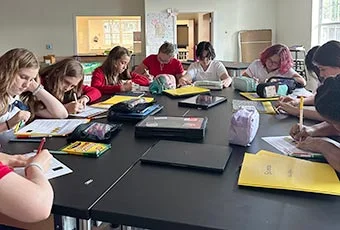Students at a large table writing and doing work together quietly.